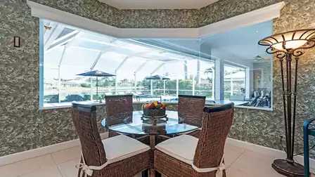 Bright dining area with round glass table, wicker chairs and large window overlooking the pool terrace and lake.